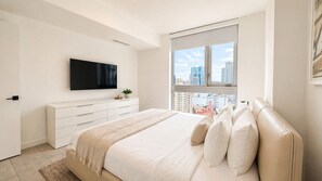 A bedroom with a bed, a TV mounted on the wall, a window with a city view, and a white dresser. - Stylish Retreat with Pool, Gym, Lounge and Private Balcony (Miami)