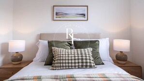 Een netjes opgemaakt bed met een geruit kussen, twee bedlampen en een ingelijst schilderij aan de muur.