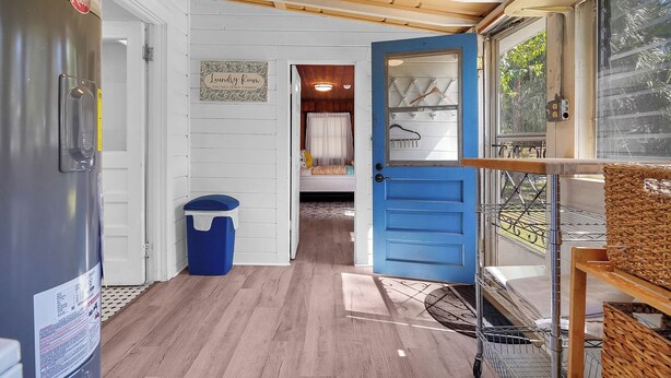 Habitació petita amb una porta blava, un frigorífic, un cistellera i un llit amb cortina blanca.