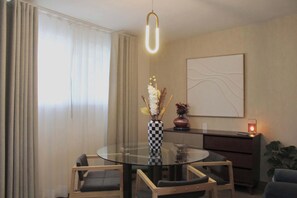 A modern dining area with a round glass table, two chairs, a vase with flowers, a wall-mounted abstract painting, and a lit candle on a side table. - MS Suite Acogedora Homeoffice Ubicadísima (Santiago de Querétaro)