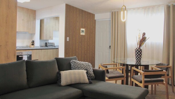 Ein modernes Wohnzimmer mit einer Eckcouch, einem Glastisch, Holzelementen und Blick auf die Küche.