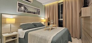 A hotel room with a bed, bedside lamps, a nightstand, a framed picture on the wall, a window with curtains, and a wooden wardrobe. - Luxury Suite Pool View at Option Near the Beach (Rio de Janeiro)