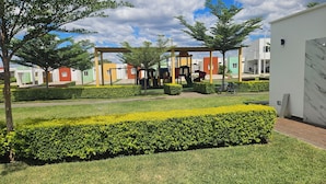 Ein gepflegter Garten mit ordentlich geschnittenen Hecken, einem Spielbereich für Kinder und modernen Gebäuden im Hintergrund.