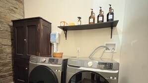 A laundry area with two front-loading washing machines, a dark wooden cabinet, and a shelf with bottles and a spray bottle. - Suite A at The Elliott 1887 (LeRoy)