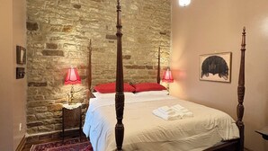 A bedroom with a stone wall, a four-poster bed, a red rug, and two bedside lamps. - Suite A at The Elliott 1887 (LeRoy)