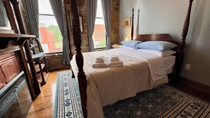 A four-poster bed with a wooden headboard, a blue patterned rug, and a view of a brick building through a window. - Suite A at The Elliott 1887 (LeRoy)