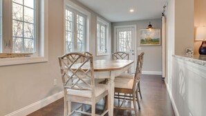 A dining area with a round table and four chairs, large windows, and a painting on the wall. - Coastal Wells Cottage with Deck and Fire Pit (Wells)
