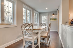A dining area with a round table and four chairs, large windows, and a painting on the wall. - Coastal Wells Cottage with Deck and Fire Pit (Wells)