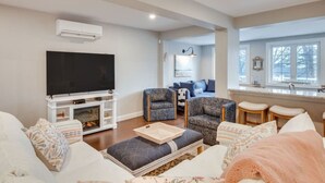 A living room with a flat-screen TV, a fireplace, a sofa, a patterned armchair, and a window with a view. - Coastal Wells Cottage with Deck and Fire Pit (Wells)