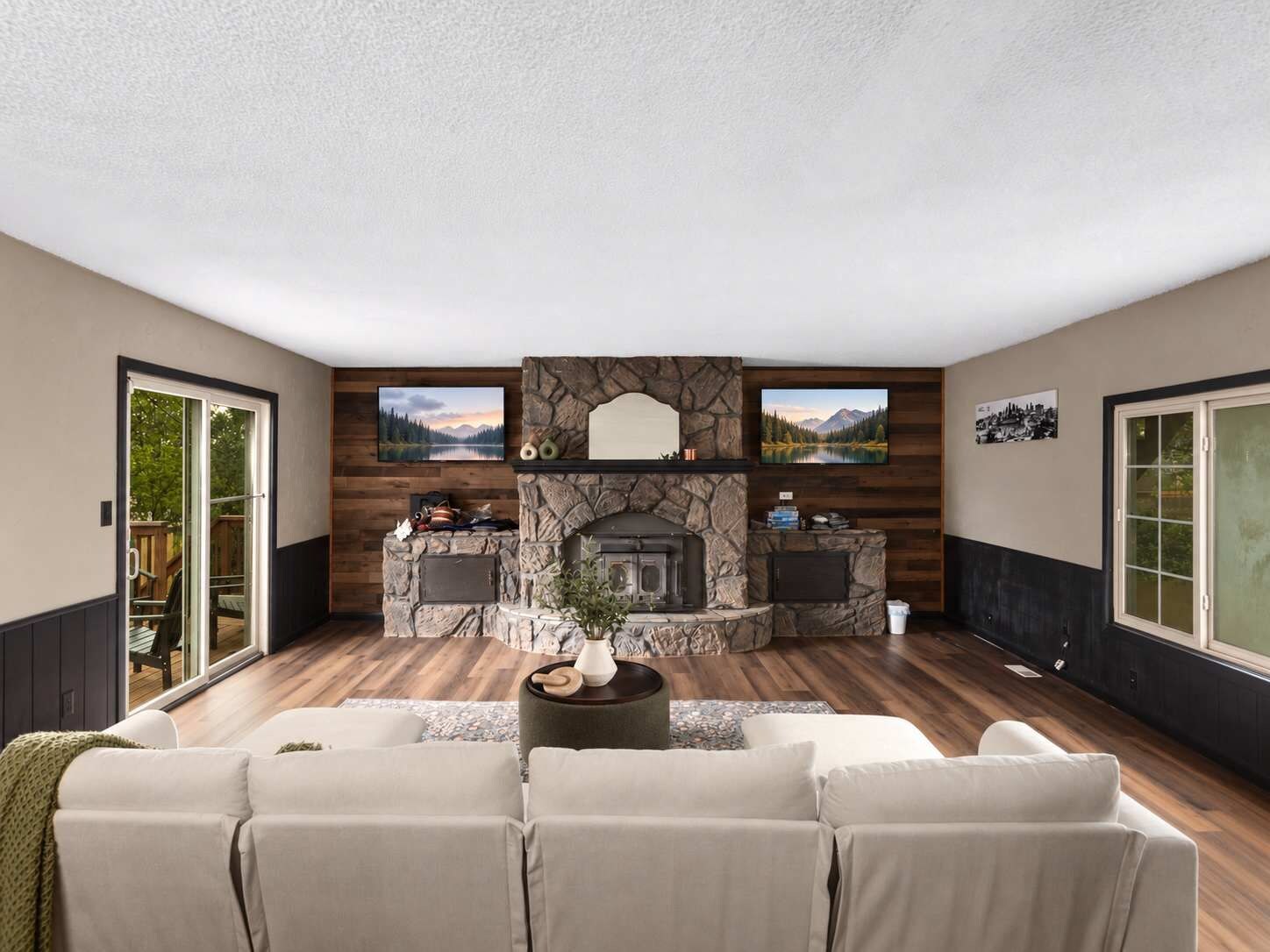 A living room with a stone fireplace, two televisions mounted on the wall, and a large window on the right.