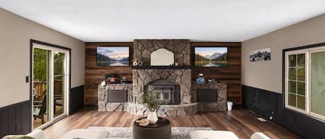 A living room with a stone fireplace, two televisions mounted on the wall, and a large window on the right.