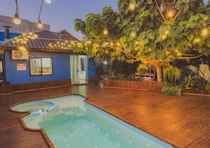 A pool area with a wooden deck, a small building with a blue exterior, and string lights. - Cabana Raízes with Hot Tub, Fireplace and Private Pool. (Chapecó)