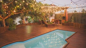 Een buitenzwembad met houten terras, omgeven door planten en een pergola met lichtslingers.