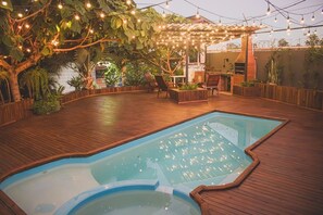 An outdoor swimming pool with a wooden deck, surrounded by plants and a pergola with string lights. - Cabana Raízes with Hot Tub, Fireplace and Private Pool. (Chapecó)