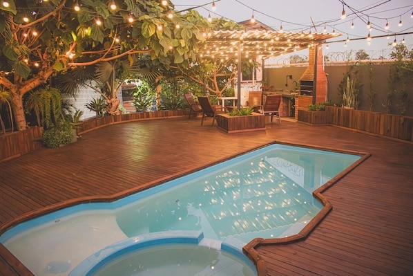 Una piscina exterior con un muelle de madera, rodeada de plantas y una pérgola con luces colgantes.
