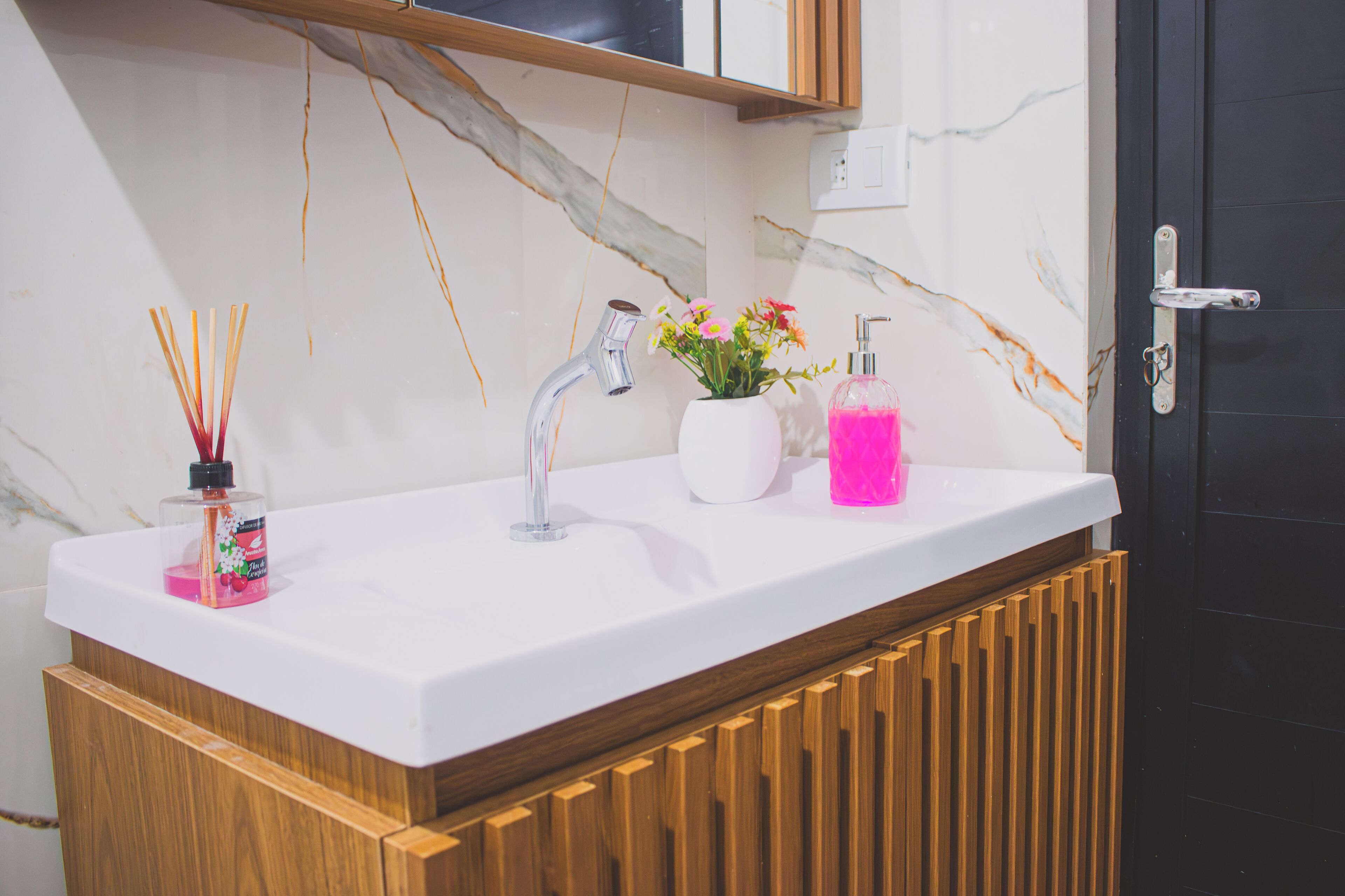 Une salle de bain avec un lavabo blanc, un meuble en bois, un mur en marbre et une porte noire.