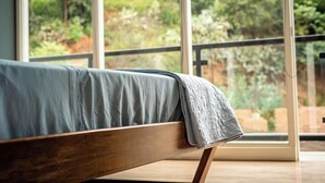 Una cama con estructura de madera y colcha azul, ubicada frente a un ventanal con vistas al exterior arbolado.