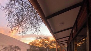 A covered outdoor area with a view of a sunset, a paved walkway, and a building with large windows. - CABAÑA CASA BLANCA HUACACHINA PERU ACCOMMODATION & TOURS (Huacachina)