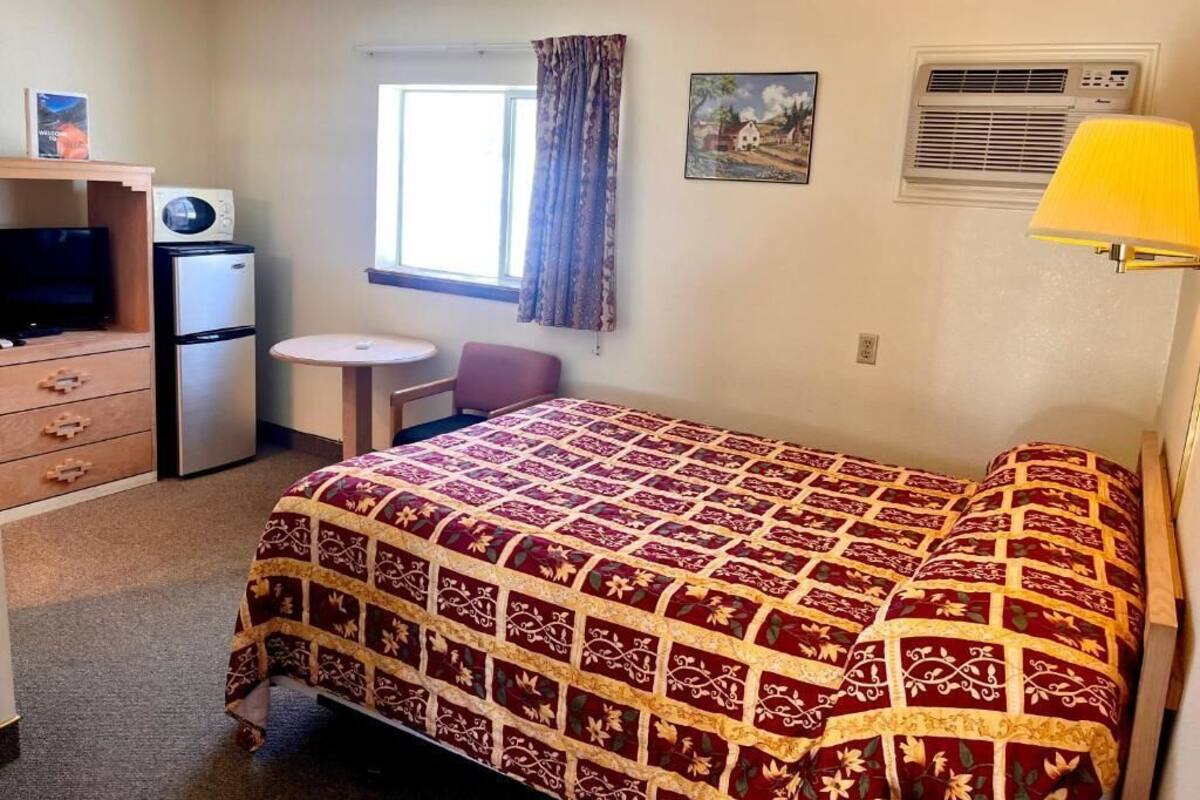 A small, well-lit room with a bed, a desk with a computer, a refrigerator, and a window with curtains.