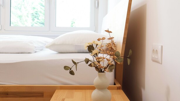 Ein Schlafzimmer mit einem Bett, einem Nachttisch aus Holz, einer weißen Vase mit Trockenpflanzen und einem Fenster mit Jalousien.