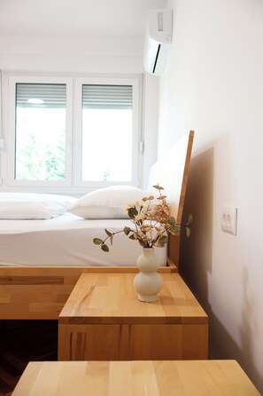 Une chambre à coucher avec un lit, une table de chevet en bois, un vase blanc contenant des plantes séchées et une fenêtre avec des stores.