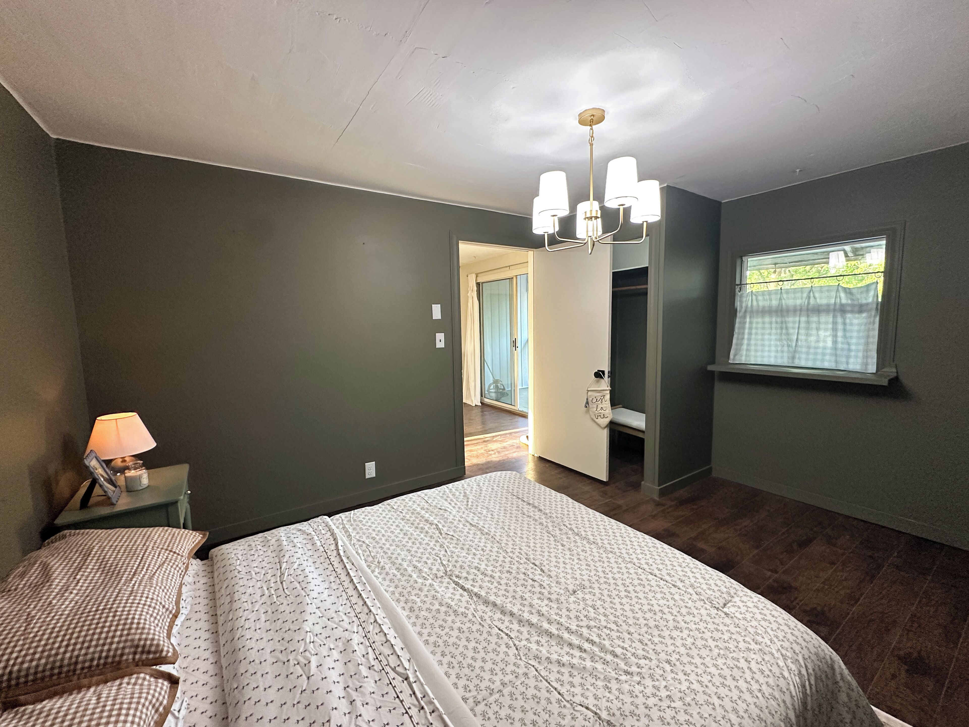 A bedroom with a bed, a nightstand, a lamp, a chandelier, a wardrobe, and a window with curtains.