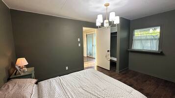 A bedroom with a bed, a nightstand, a lamp, a chandelier, a wardrobe, and a window with curtains.