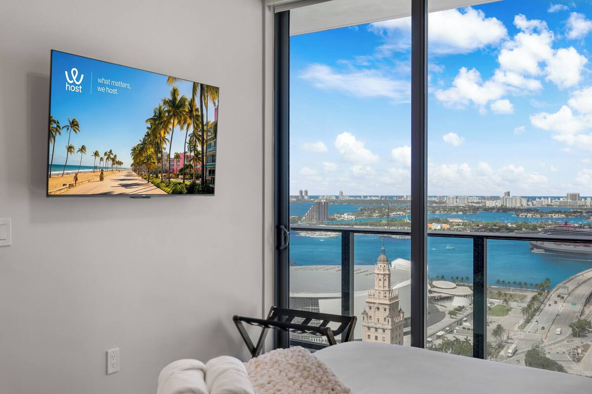 A hotel room with a large window offering a city view, a flat-screen TV on the wall, and a bed with a white blanket.