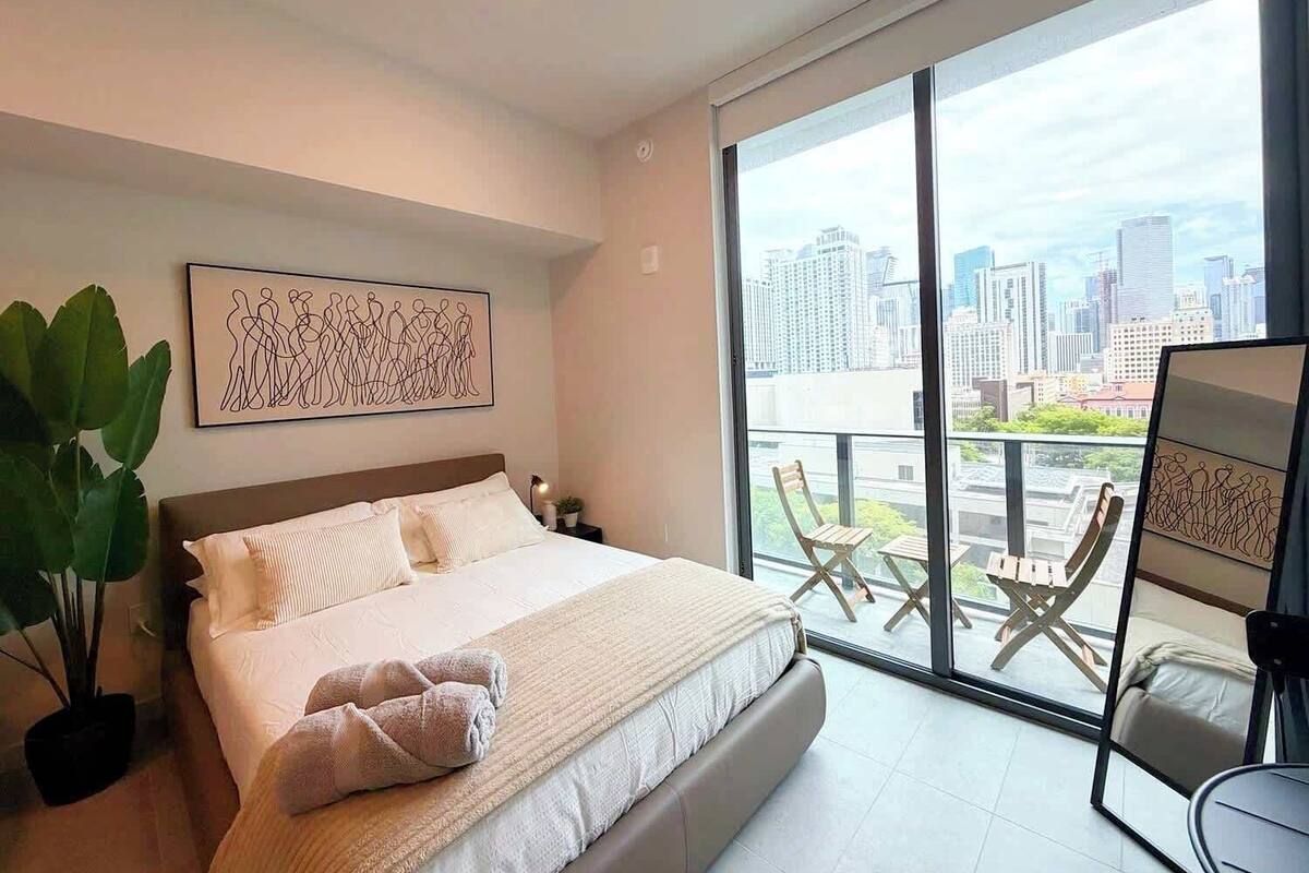 A modern hotel room with a large bed, a city view, a balcony with chairs, and a large plant.