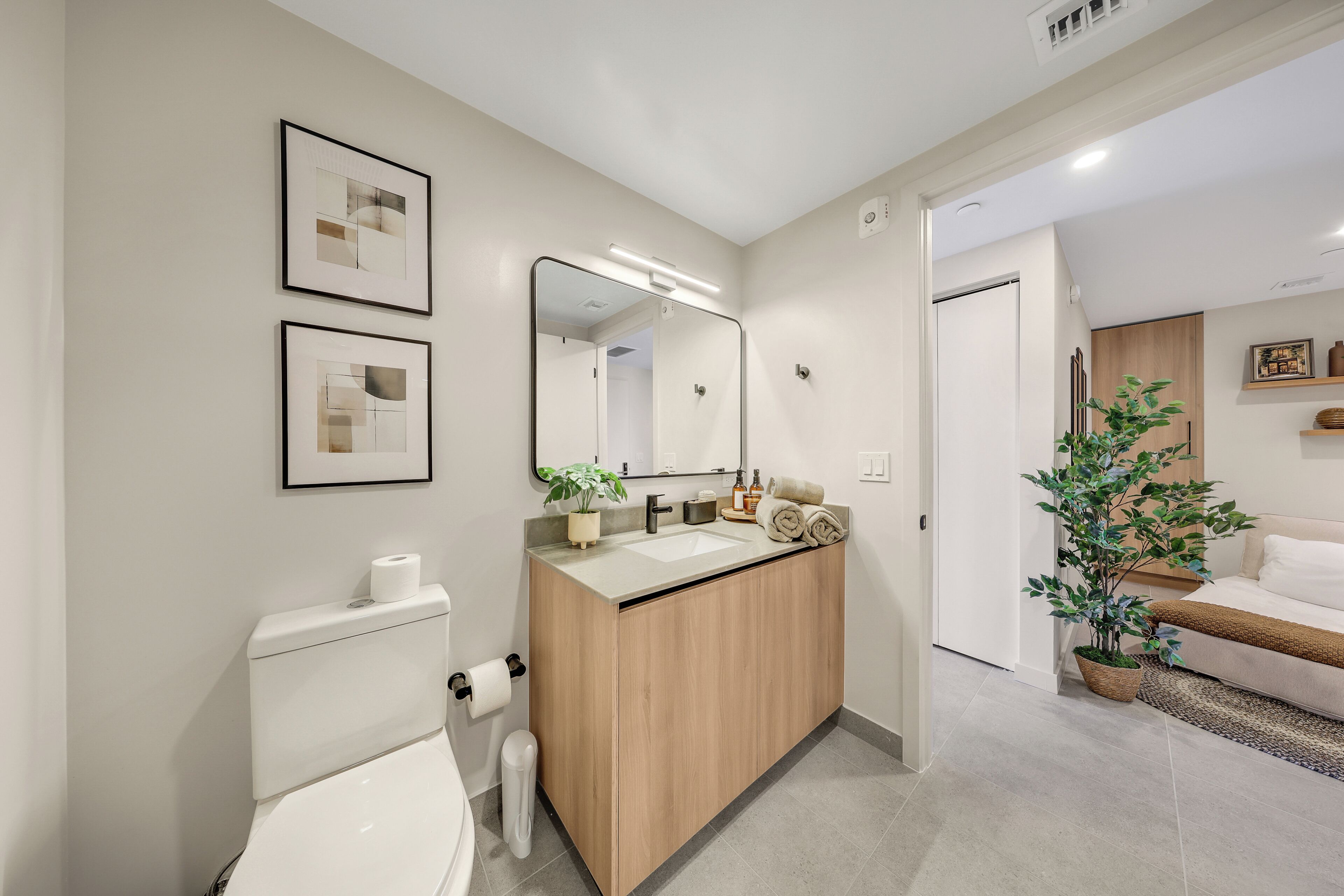 Un baño moderno con inodoro, lavamanos y espejo, conectado a una sala de estar con sofá y planta en maceta.