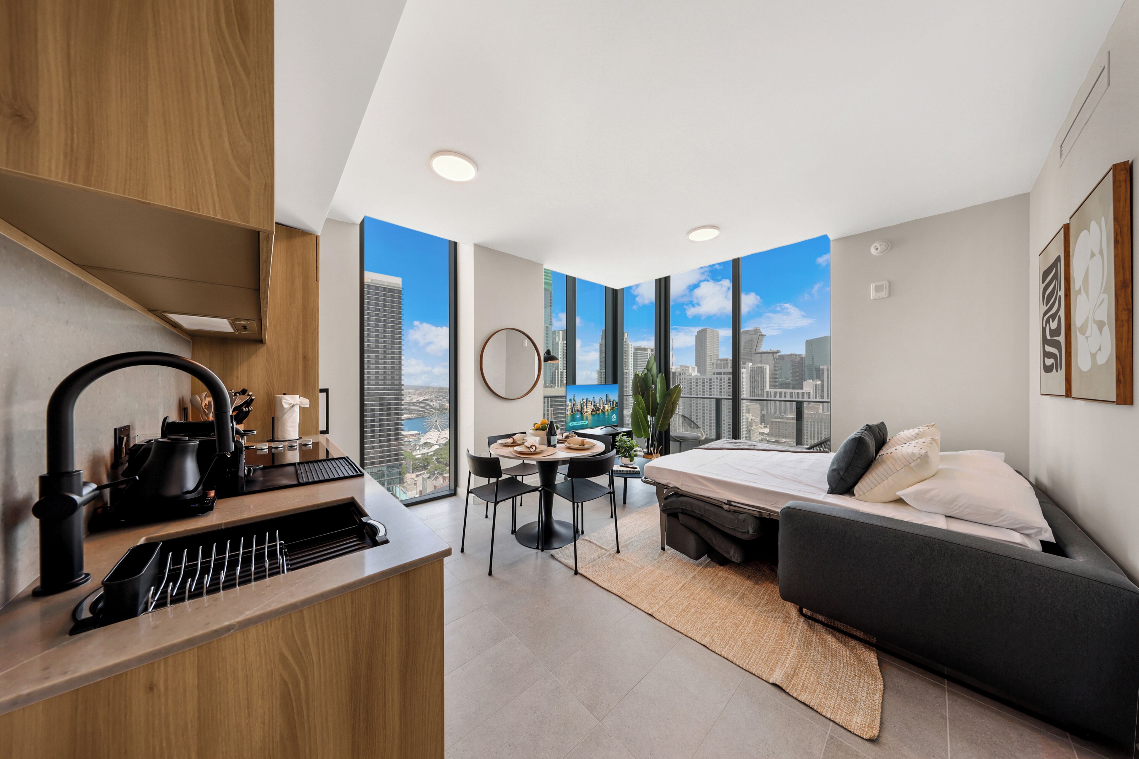 A modern hotel room with a kitchenette, a dining area, a bed, and a large window offering a city view.