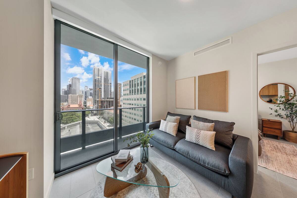 A modern living room with a large window, a glass coffee table, a grey sofa, and a city view.