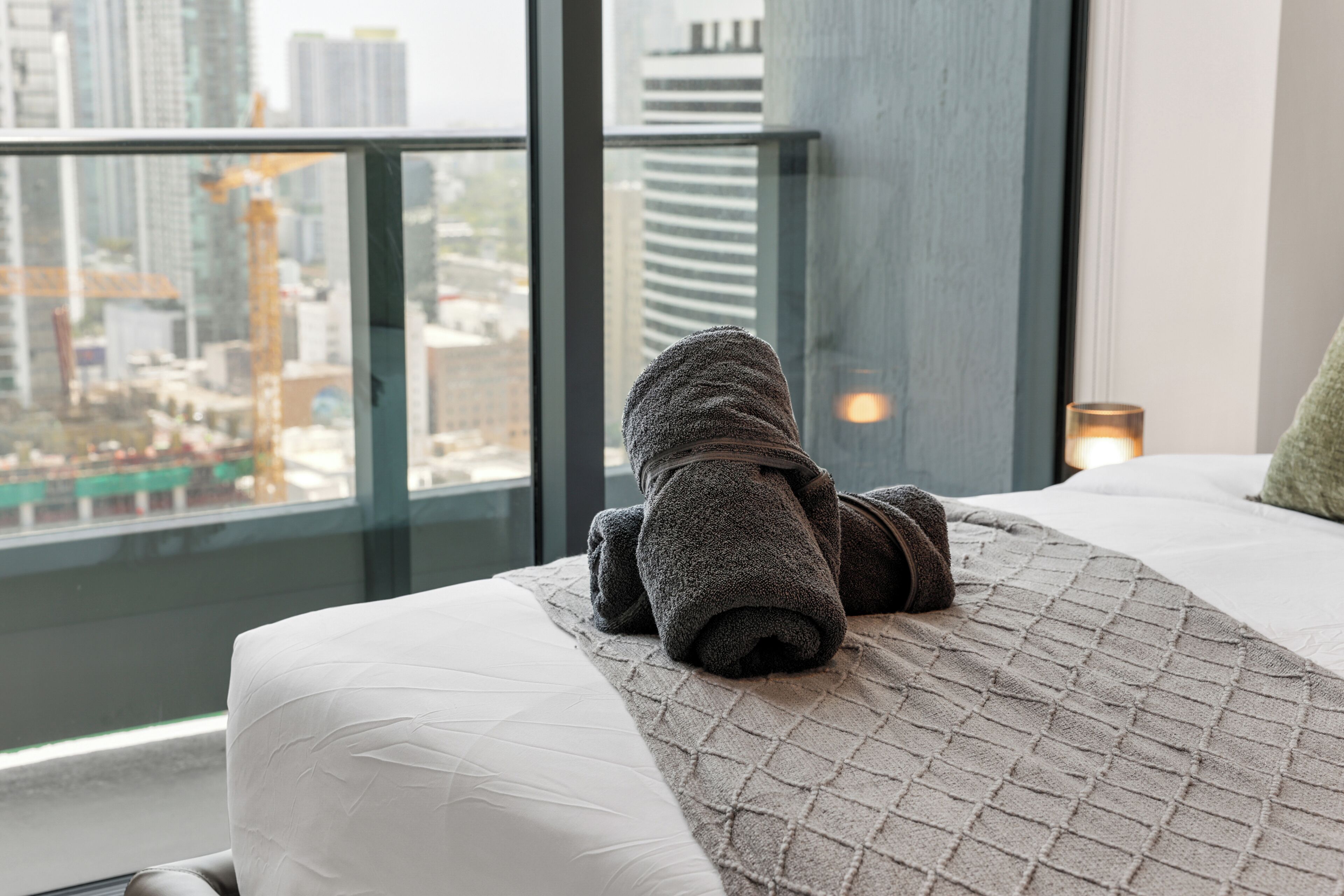A hotel room with a bed, a view of a cityscape through large windows, and a nightstand with a lamp.