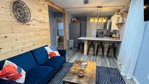 Una sala de estar con un sofá azul, una mesa de centro y un tapete estampado. Hay una zona de cocina con taburetes de bar y un ventilador de techo.
