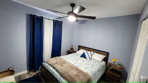 Un dormitorio con cama, ventilador de techo, mesita de noche y ventana con cortinas.