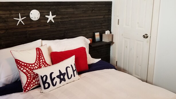 Un dormitorio con cama, cabecera, decoración en la pared, una puerta y mesita de noche con lámpara.