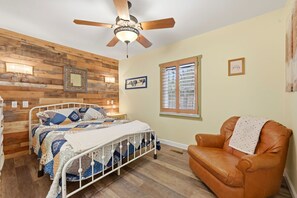 Ein Schlafzimmer mit Bett, Sessel, Deckenventilator, Fenster mit Rollläden und einer Holzwand.