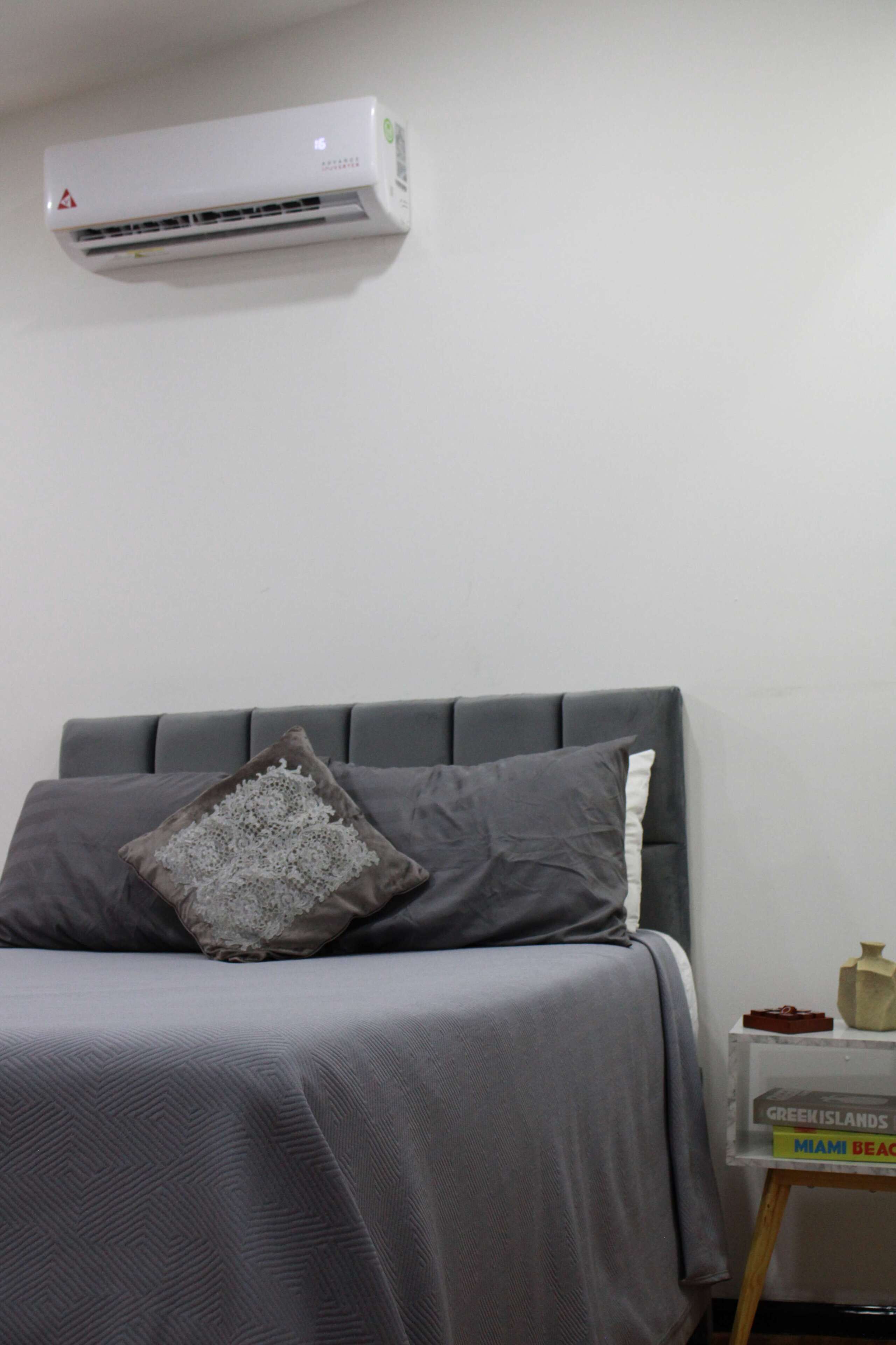 A bedroom with a bed, a pillow, a bedside table with a book, and an air conditioner on the wall.