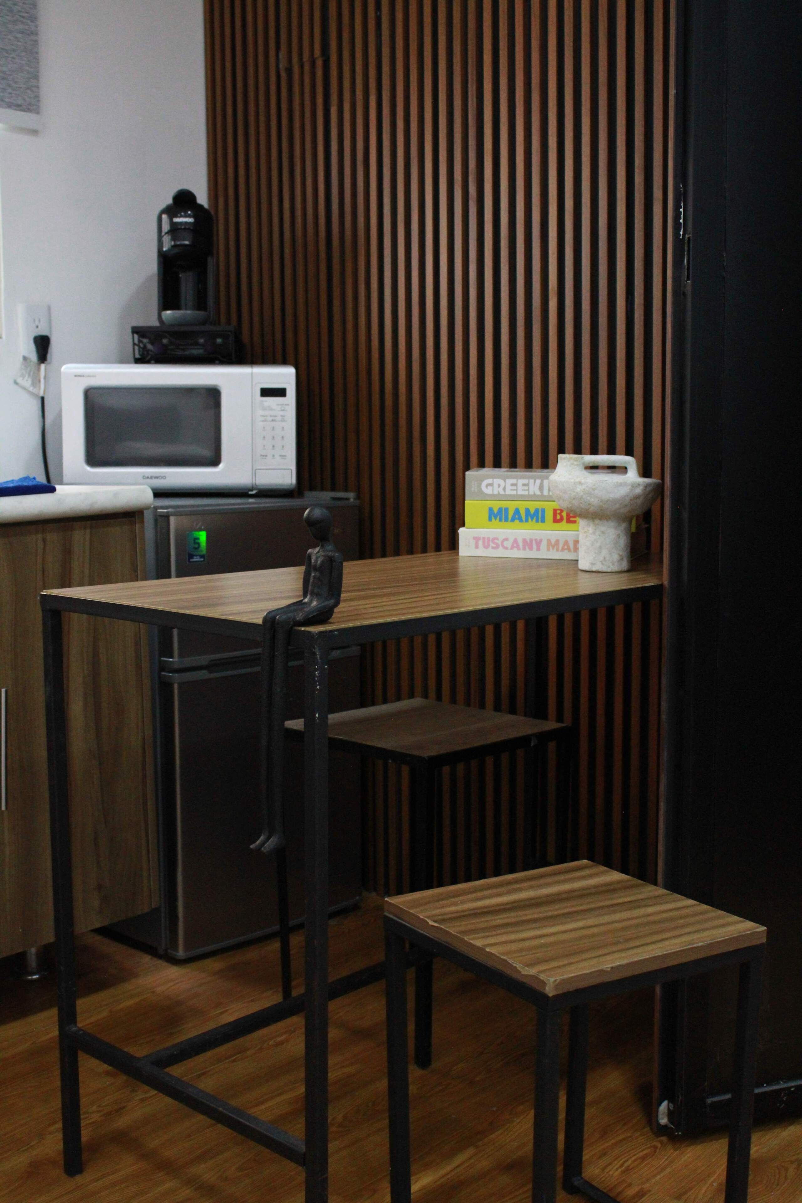 Uma kitchenette compacta com micro-ondas, cafeteira e um pequeno frigorífico. Há uma mesa de madeira com uma cadeira e um apoio para os pés.