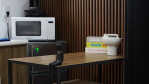 Une petite cuisine équipée d’un four à micro-ondes, d’une machine à café et d’un petit réfrigérateur. On y trouve une table en bois avec une chaise et un petit tabouret.