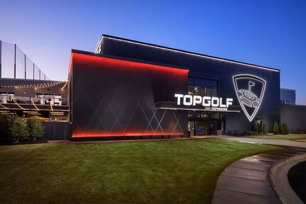 Un bâtiment moderne arborant un logo TOPGOLF bien visible, une pelouse bien entretenue et un ciel dégagé.