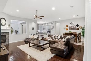 A modern living room with a fireplace, a ceiling fan, and a dining area with a kitchen in the background.