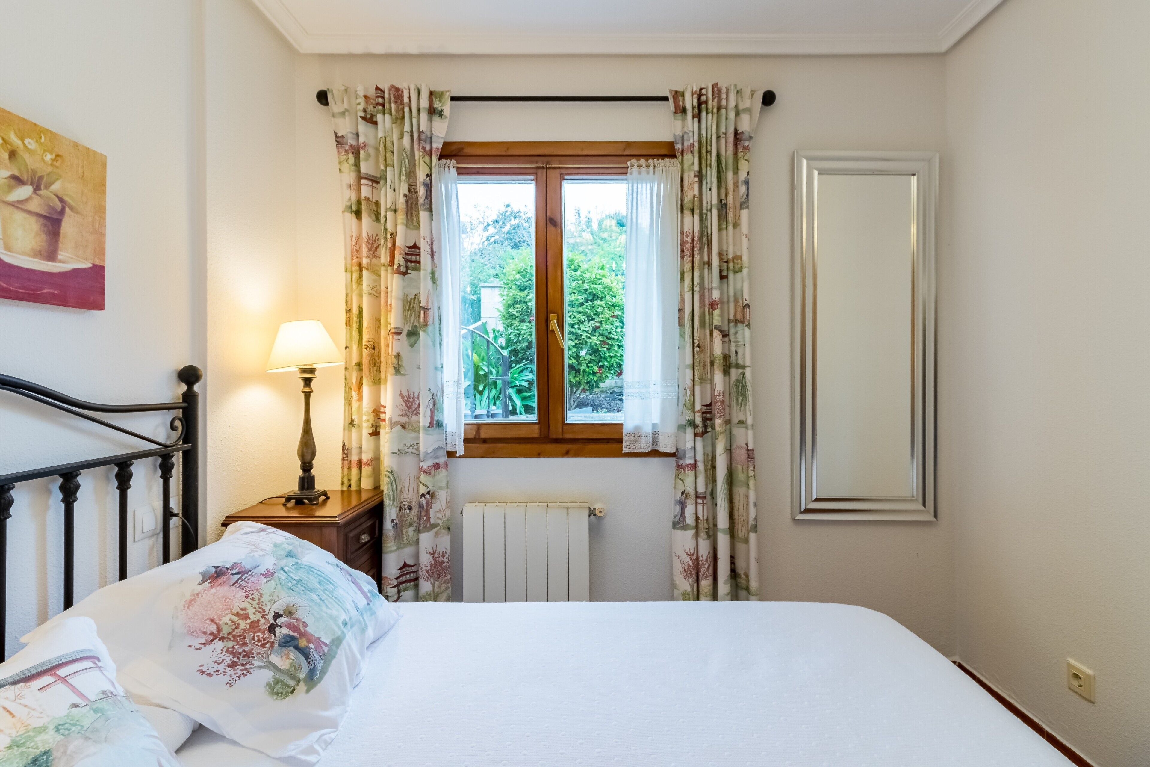 A bedroom with a bed, a nightstand with a lamp, a window with curtains, and a radiator.