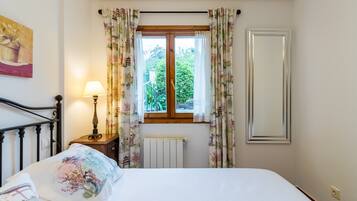 A bedroom with a bed, a nightstand with a lamp, a window with curtains, and a radiator.