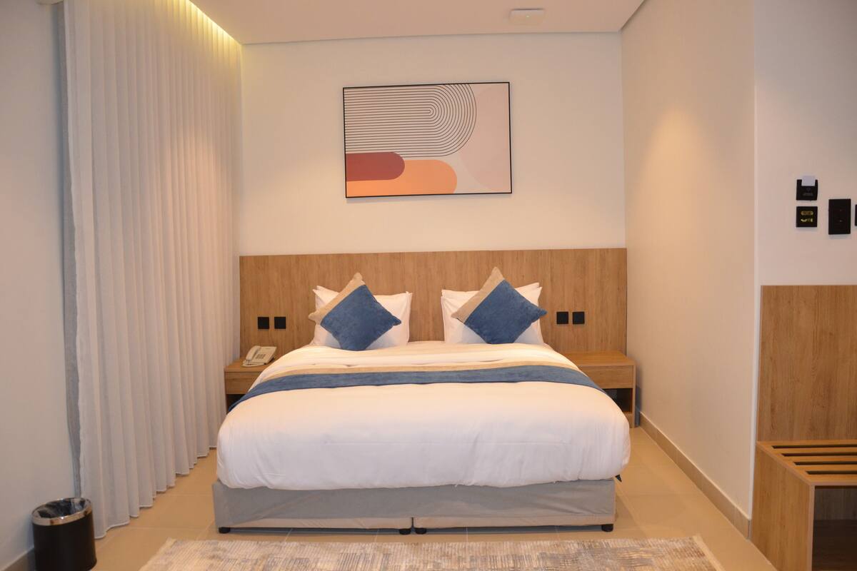 A modern hotel room with a large bed, wooden headboard, two bedside tables, a telephone, and a wall-mounted artwork.
