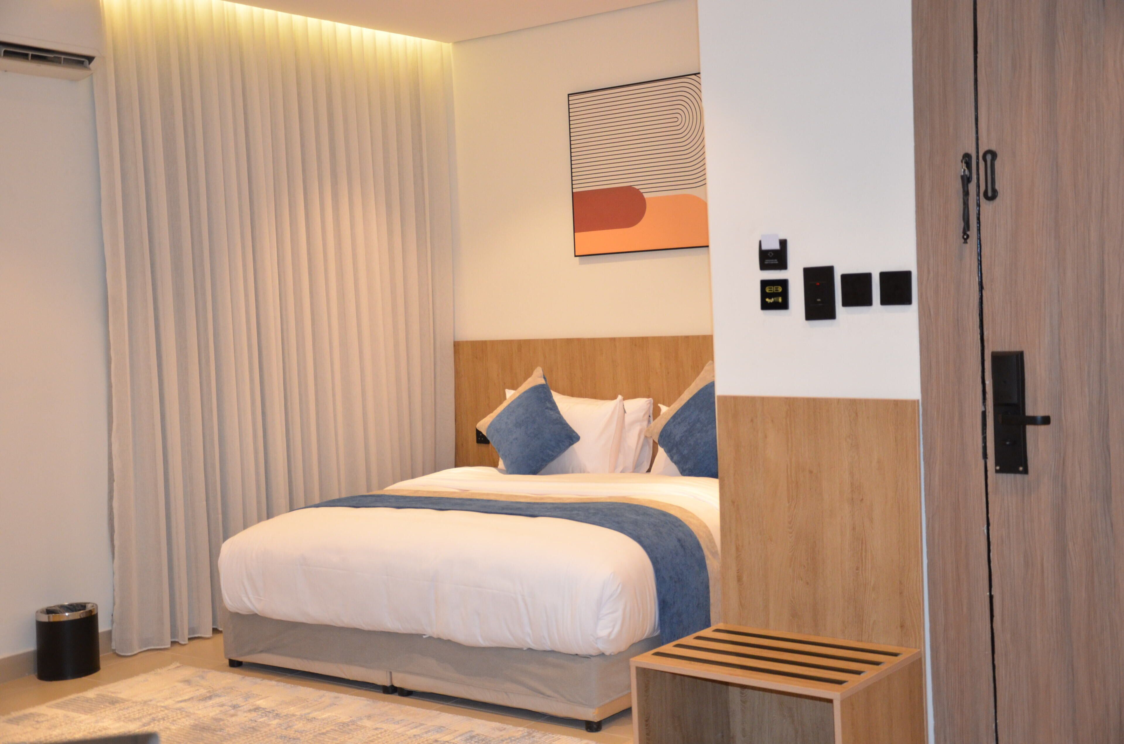 A modern hotel room with a large bed, a wooden headboard, a bedside table, a wall-mounted artwork, and a wardrobe.