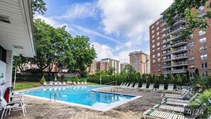 Una piscina con sillas de descanso, rodeada de árboles y edificios altos.
