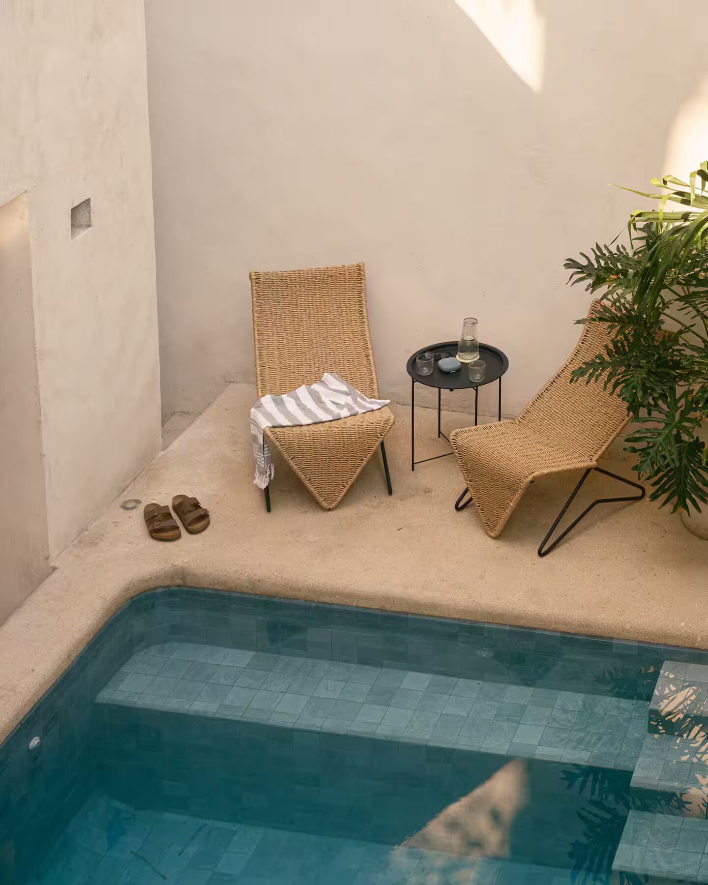 Ein kleiner Poolbereich im Freien mit zwei Korbstühlen, einem Tisch mit einem Glas und einer Kerze sowie einem Paar Sandalen am Boden.