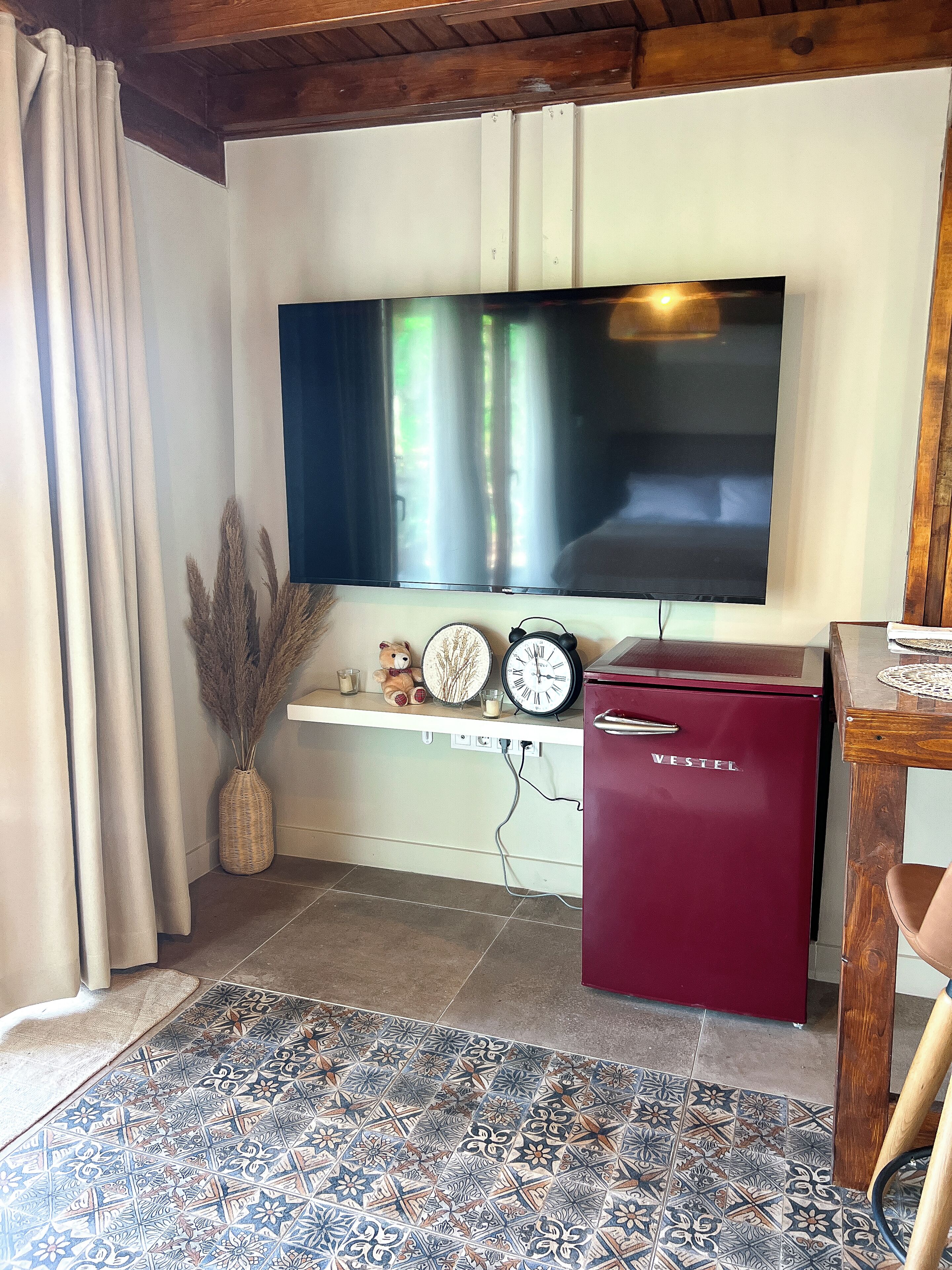 Una sala de estar con un televisor de pantalla plana montado en la pared, un refrigerador VESTEL rojo, un reloj, un jarrón con plantas secas y una alfombra estampada.