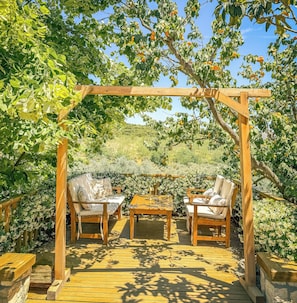 An outdoor wooden pergola with a table and two chairs underneath, surrounded by lush greenery and fruit trees. - Casa Del Olivia (Seferihisar)